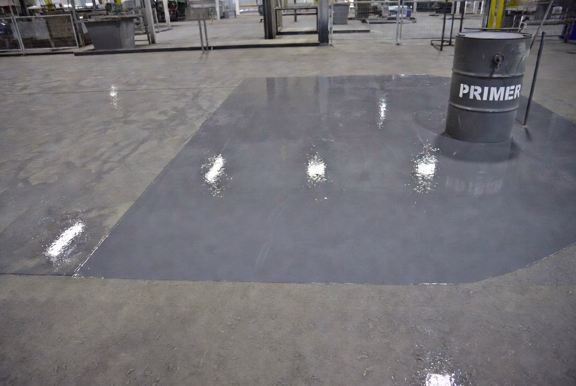 Endüstriyel beton zeminde primer uygulanmış yüzey bölümü ve primer varili